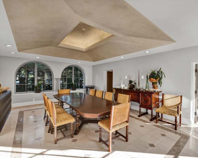A spacious dining room features a large wooden table surrounded by upholstered chairs and windows that overlook a garden.