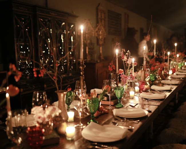 A long wooden dining table is elegantly set with candles, decorative flowers, and glassware in a dimly lit room.