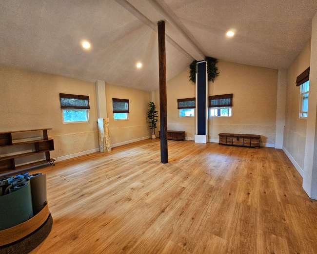 A spacious and well-lit room features wooden floors, large windows, and minimal furnishings, including yoga mats and benches.