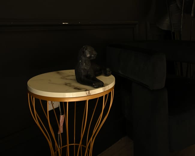 A black decorative figurine of an animal sits on a marble-topped table with a gold wire base, against a dark wall and a plush black sofa.