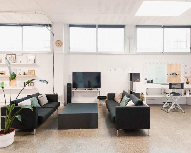 An open studio space with two black sofas facing a coffee table, a large television, and a mirror setup in a well-lit room with large windows.