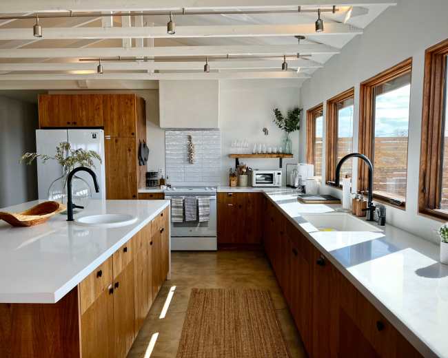 A modern kitchen features wooden cabinetry, white countertops, and ample natural light from large windows.