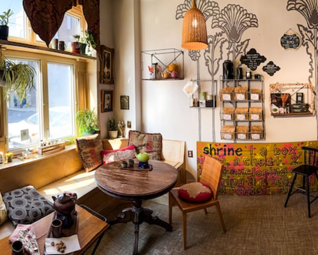 The image depicts a cozy café interior with various seating options, light wood furniture, and a decorative wall featuring a tree mural.