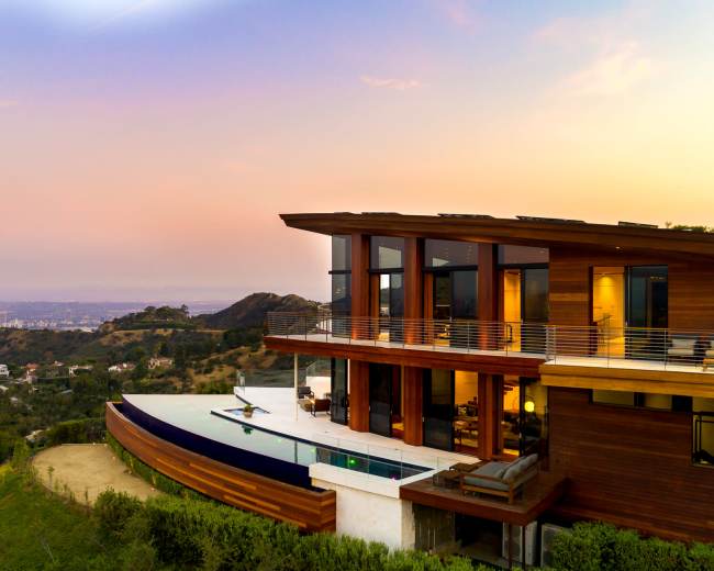 A modern house with large windows overlooks a city skyline at sunset, set against a hillside.