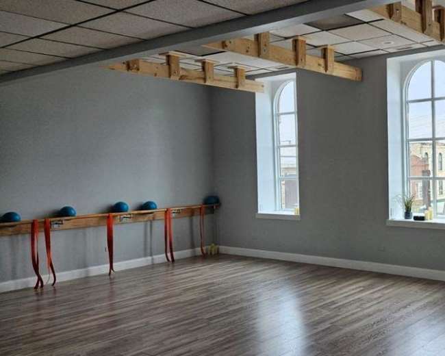 The image shows a spacious, empty room with large windows and wooden beams overhead, featuring equipment lined up against one wall.