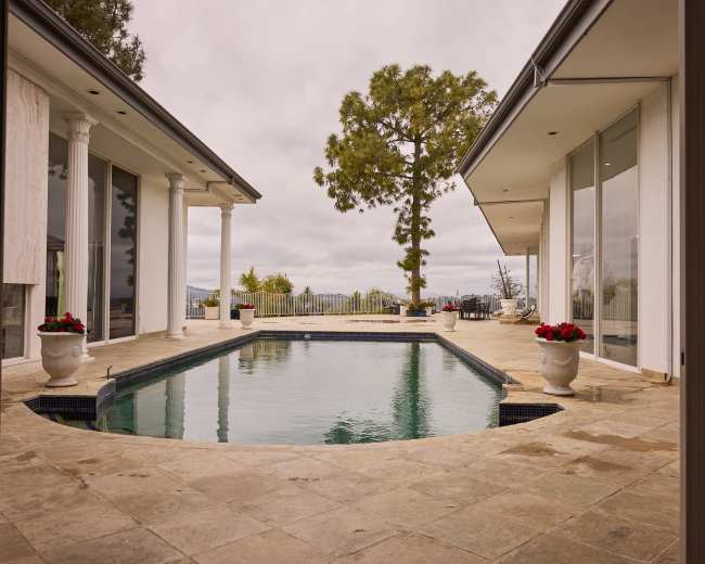 A spacious modern home features an infinity pool surrounded by stone tiles and large windows with views of distant hills and trees.