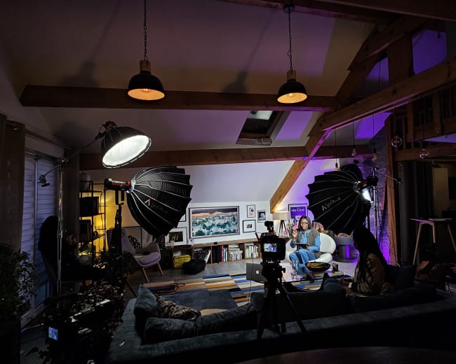 A well-lit indoor scene features two people seated on a couch in a stylish living room, surrounded by professional photography equipment and colorful lighting.