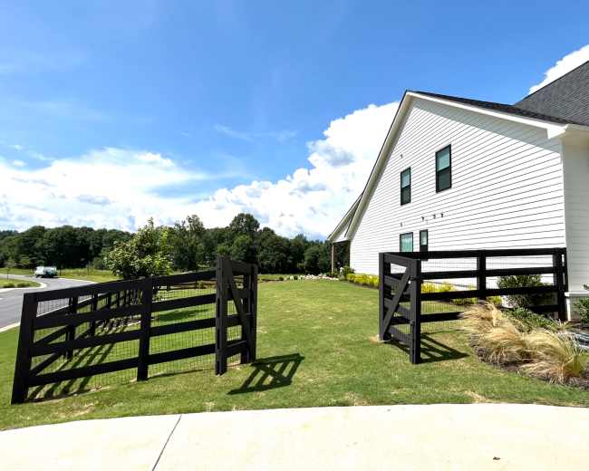 A black wooden fence with two gates leads to a grassy area beside a white building under a blue sky.