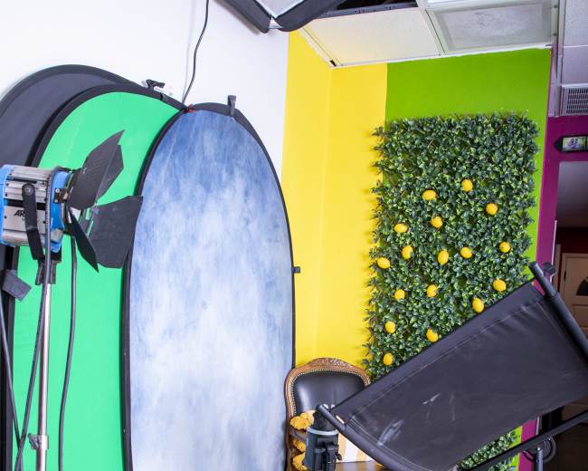 A photography studio setup featuring a green screen, a backdrop, lighting equipment, and a chair placed in front of a decorative wall with faux greenery and yellow accents.