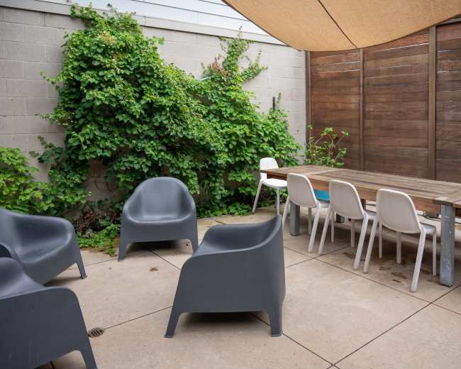 A patio area features a wooden table surrounded by chairs and shaded by a large canopy, with greenery climbing a nearby wall.