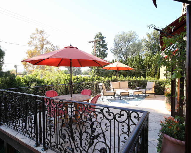 A patio features a wooden table surrounded by red chairs and two large red umbrellas overlooking a landscaped yard.