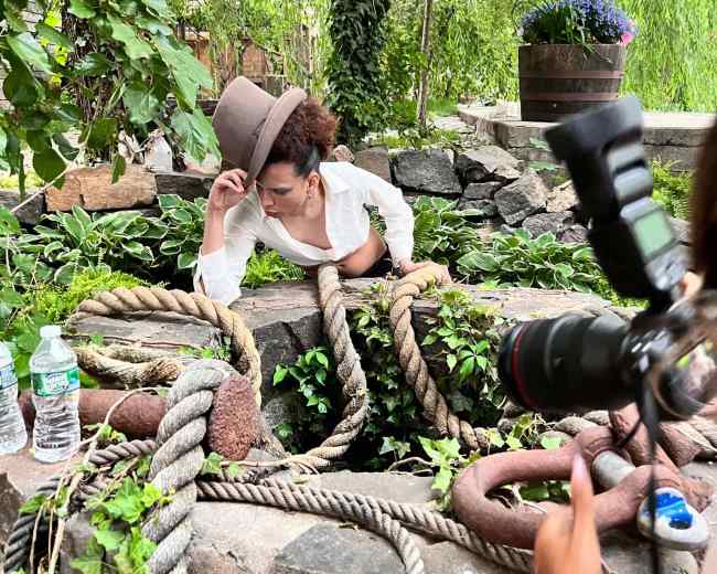 A person is emerging from a stone well surrounded by greenery and old ropes, while another individual nearby is holding a camera.