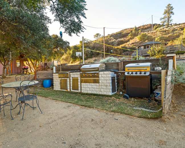 A wooden barn with a rustic aesthetic stands beside a grassy area, featuring a dog lying in front and hay bales nearby.