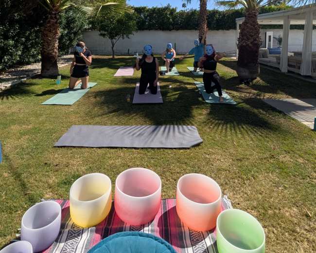 A group of people practices yoga on mats in a grassy area surrounded by palm trees, with colorful sound bowls arranged in the foreground.