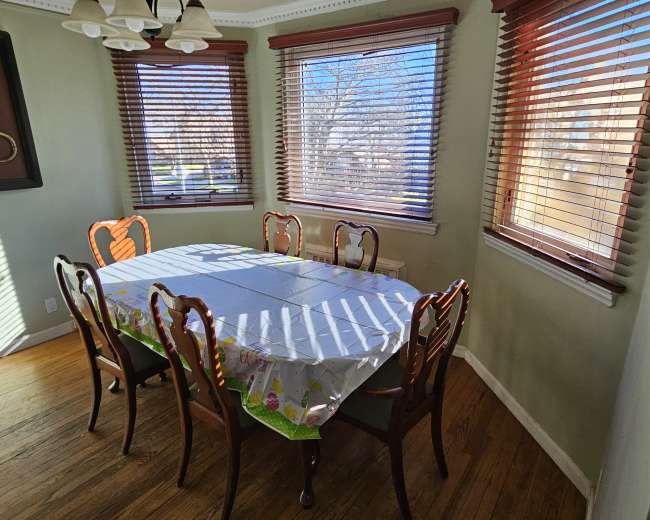 A dining table with six chairs is set in a well-lit room featuring window blinds and hardwood flooring.