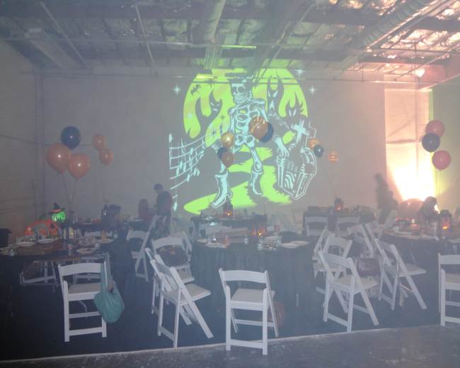 A Halloween-themed party scene features tables and chairs arranged around a space with a colorful skeleton projection on the wall, along with orange and black balloons.