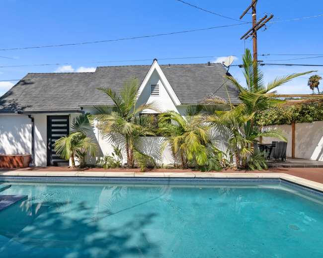 A modern white house is situated beside a clear blue swimming pool, surrounded by palm trees and a fenced patio area.