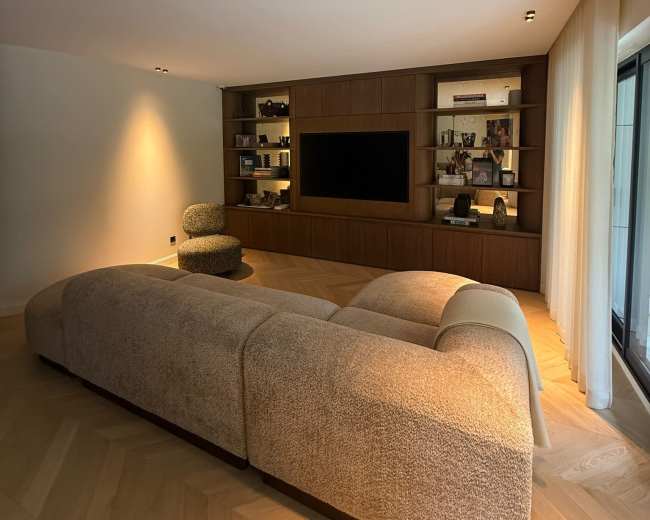 The image shows a modern living room with a curved sofa, a wall-mounted television, and wooden shelving units displaying decorative items.