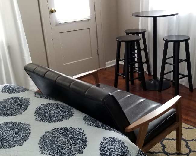 A small room with a bed in the foreground and a black table with three stools in the background, next to a chest of drawers.