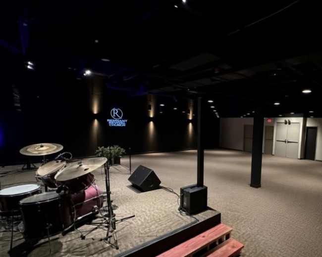 The image shows a dimly lit event space with a stage for musical performances on the left and an empty floor area on the right.