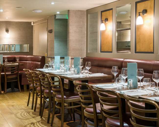 The image shows a restaurant interior featuring several tables set with glasses and utensils, surrounded by red leather seating and wooden flooring.