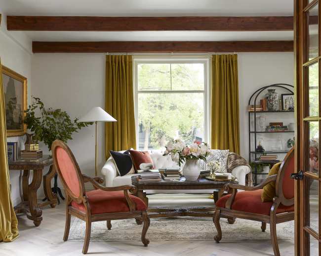 The image shows a living room featuring a white sofa, two red-upholstered armchairs, a wooden coffee table, and large windows with yellow curtains, framed by a set of double doors.