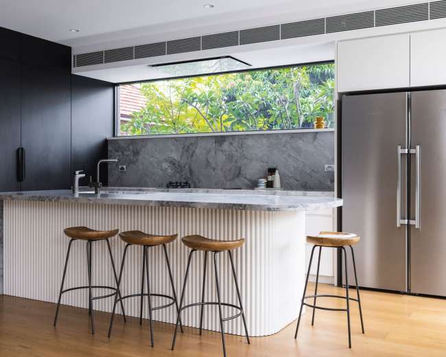 A modern kitchen features a marble-topped island with four wooden stools, stainless steel appliances, and large windows overlooking greenery.