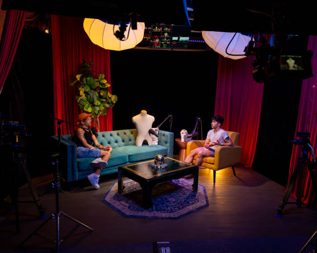 A talk show set features two people seated on a teal sofa and a beige chair, with cameras and lighting equipment positioned around them.