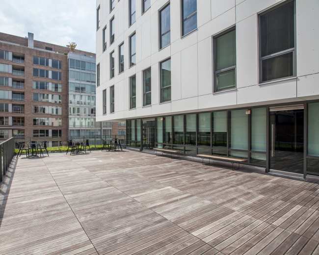 The image shows a modern outdoor terrace with wooden flooring adjacent to a building with large glass windows.