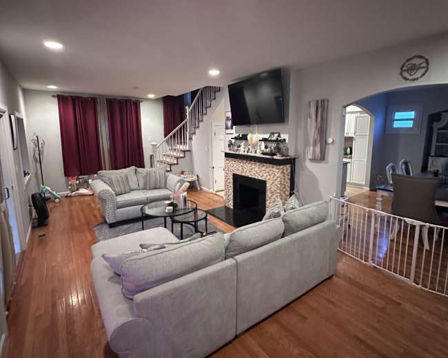 A spacious living room with gray sofas, a fireplace, a large television mounted on the wall, and a staircase leading upstairs.