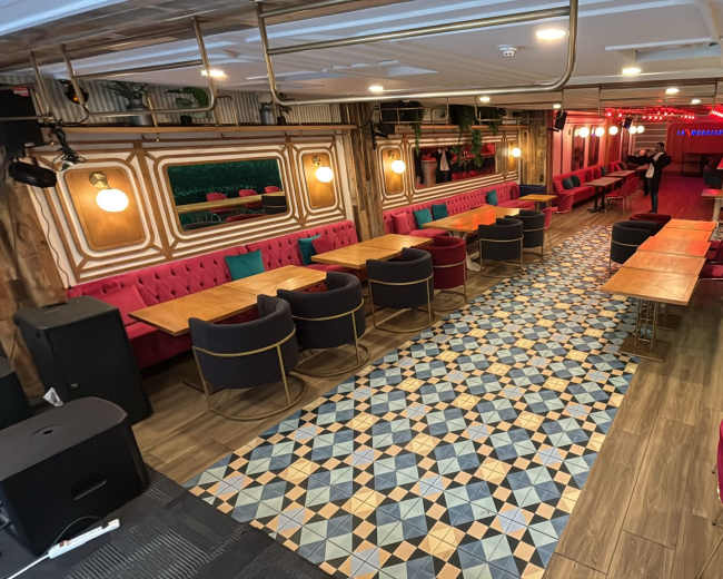 The image shows a spacious restaurant interior with wooden tables, colorful chairs, and patterned flooring, illuminated by warm lighting.