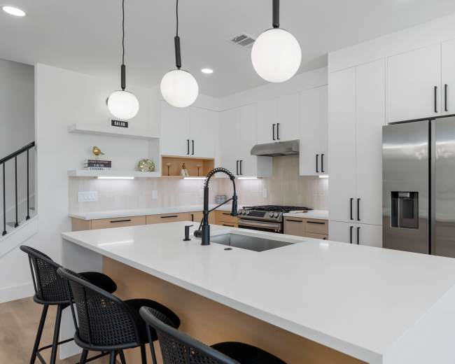The image shows a modern kitchen featuring a large island with bar seating, a stainless steel refrigerator, and pendant lighting above the countertop.