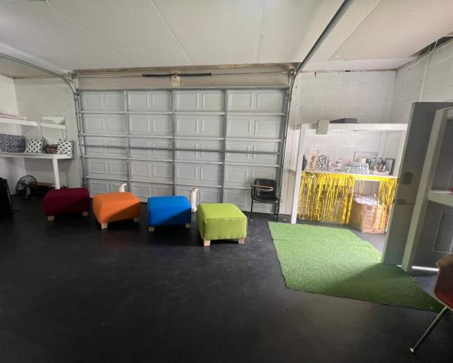 The image shows a garage-like space featuring colorful ottomans arranged in a row, with a kitchenette and a grass-like mat in the corner.