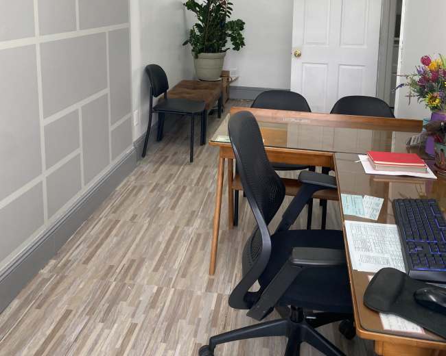 A small, tidy office space with a wooden desk, several black chairs, and a potted plant in the corner.