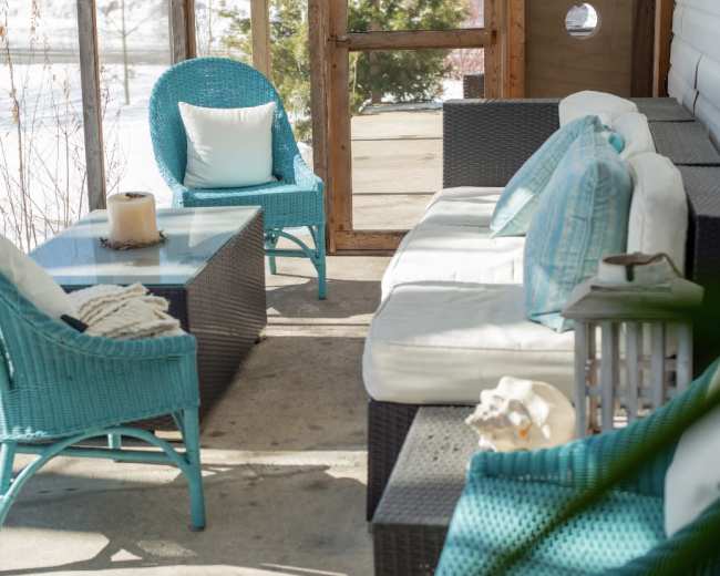 A sunlit porch features a glass table surrounded by turquoise wicker chairs and a gray wicker sofa.