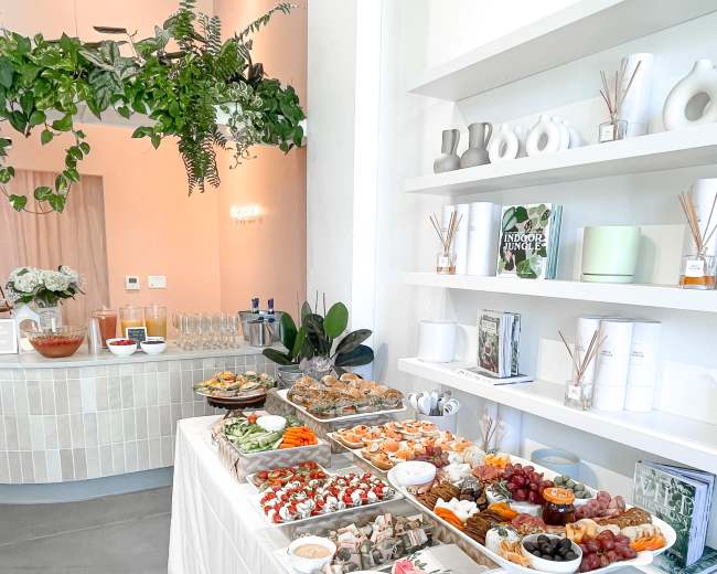 A white tablecloth displays a variety of food platters, including seafood, fruits, and pastries, against a backdrop of decorative shelves and greenery.