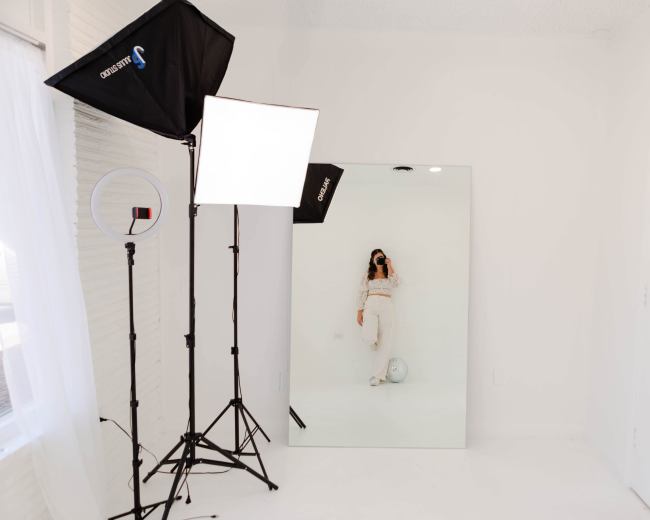 A person stands in front of a large mirror in a brightly lit, minimalist room with studio lighting equipment.