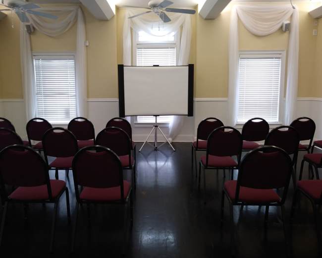 A meeting room is set up with rows of red chairs facing a blank projection screen on a tripod between two windows.