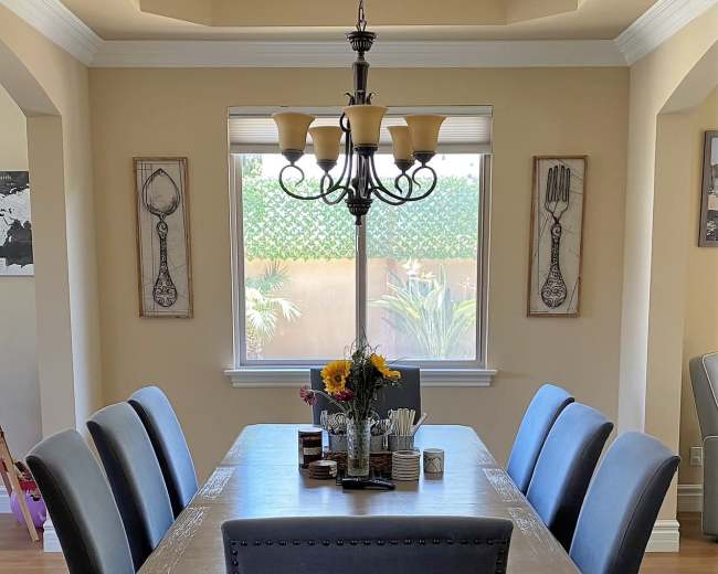 A dining room features a long table surrounded by six gray chairs, with a chandelier hanging above and a vase of sunflowers in the center.