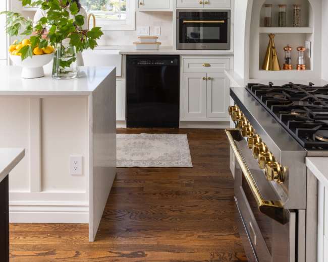 A modern kitchen features white cabinetry, a black dishwasher, a gas stove with brass accents, and a vase of flowers on the countertop.
