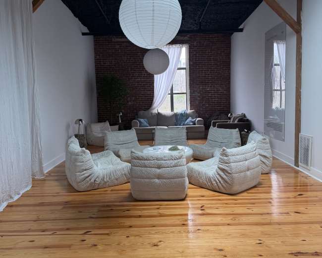 A spacious living area features a circular arrangement of cushioned seating, wooden floors, and large windows with natural light.