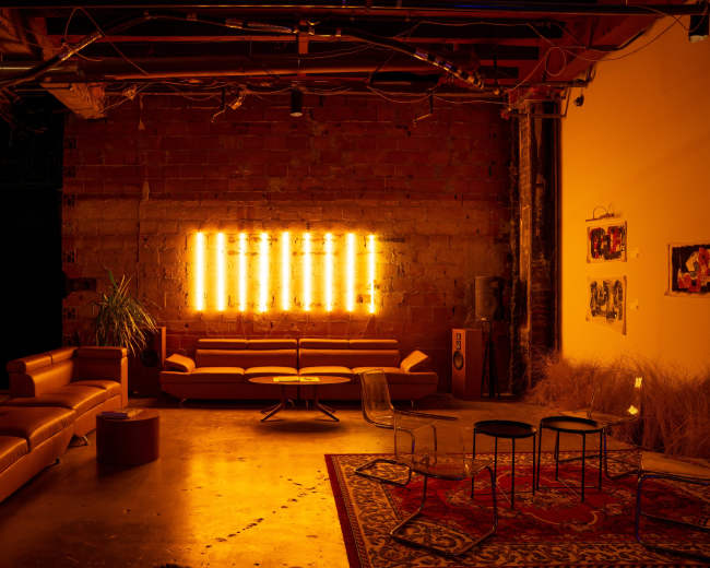 The image shows a dimly lit lounge area with orange neon lights on the wall, leather sofas, a coffee table, and a patterned rug.