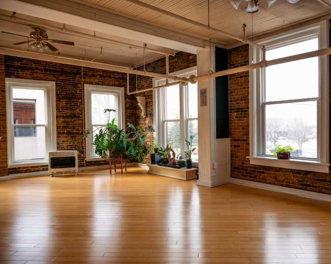 The image shows a spacious yoga studio with exposed brick walls, large windows, and wooden flooring.