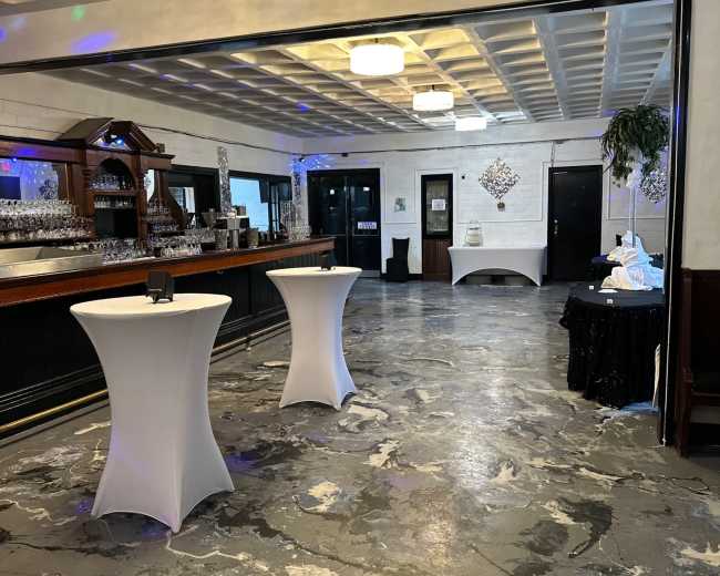 An empty event space with a bar area, white cocktail tables, and a polished concrete floor.