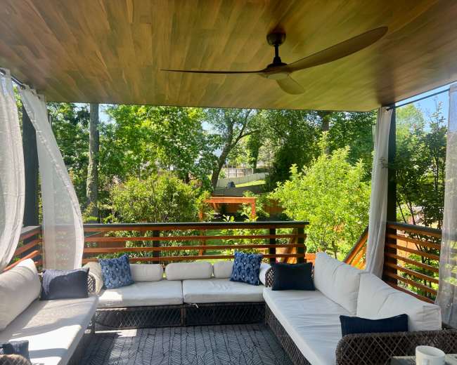 A covered outdoor seating area with a sectional sofa, decorative pillows, and a fan, surrounded by greenery.