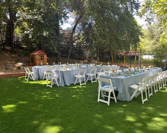 A large outdoor dining setup with long tables and white chairs is arranged on green turf, surrounded by trees and children's play equipment.