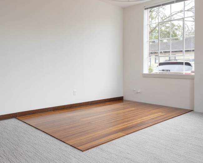 The image shows a sparsely furnished room featuring a section of hardwood flooring and a large window.