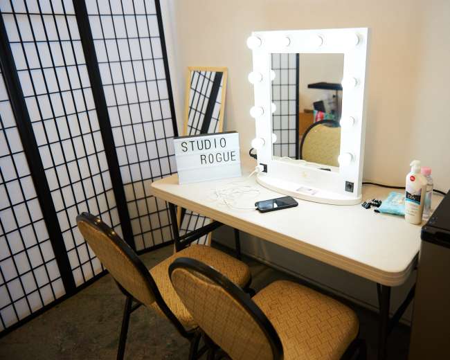A well-lit vanity mirror with bulbs is positioned on a table alongside a sign that reads "STUDIO ROGUE," two chairs, and a small refrigerator in a room with partition screens.