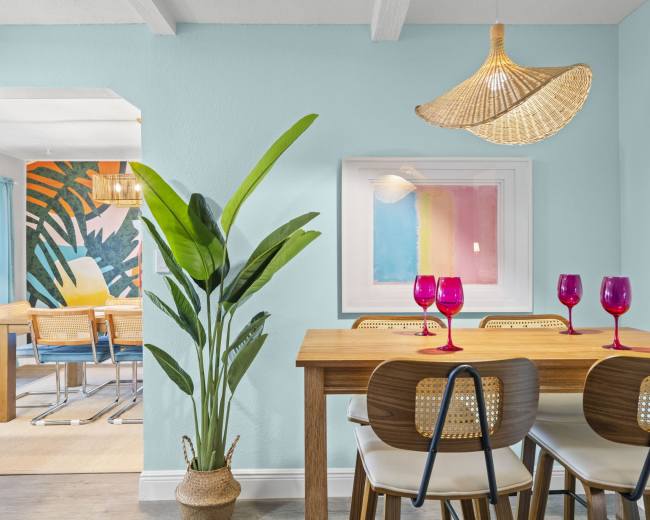 A dining area features a wooden table set with colorful glasses, surrounded by white chairs and a potted plant, with an art piece and an adjacent room visible in the background.