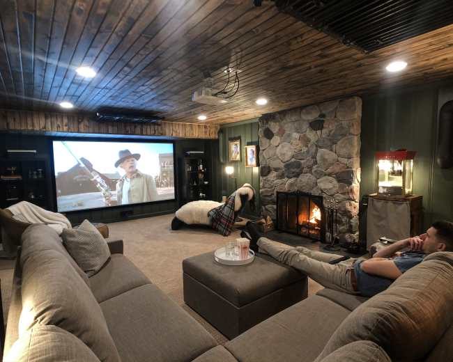 A person relaxes on a grey sectional sofa while watching a movie on a large screen in a cozy, stone-walled basement with a fireplace.